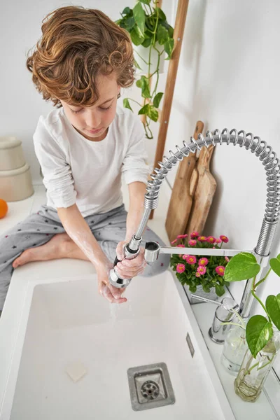 朝のシンクで洗浄しながら 石鹸の手に蛇口から水を注ぐパジャマで裸足の巻き毛の少年の高い角度 — ストック写真