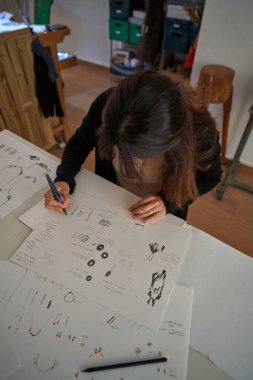 Yüzü olmayan kadın tasarımcının kuyumcudaki kağıt tabaka üzerinde yaratıcı el yapımı aksesuarlar çizdiği yüksek açı.