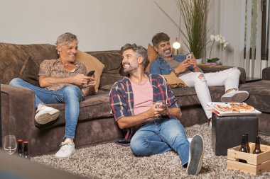 Hafta sonu partisinde rahat oturma odasında oturan akıllı telefonları kullanan gülümseyen erkek arkadaşlar.