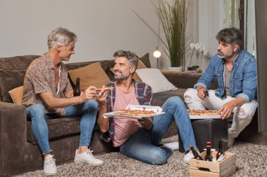 Mutlu olgun erkekler, günlük kıyafetlerini giyip evde eğlenirken lezzetli pizzalarını yiyorlar.