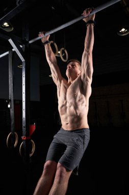 Üzerinde siyah arka plan olan spor salonunda barfiks çeken konsantre, güçlü, üstsüz erkek vücut geliştirici.