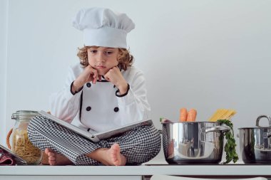 Aşçı kıyafetli, şapkalı, çapraz bacaklı ve havuç ve spagettiyle tencere kenarında yemek kitabı açan düşünceli reşit olmayan çocuktan.