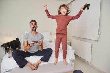 Uzun boylu, duygusal bir adam ve neşeyle zıplayan bir çocuk köpekle yatakta video oyunu oynuyorlar.