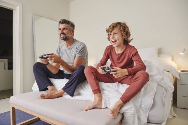 Uzun boylu, çıplak ayaklı baba ve oğlu video oyunu oynarken yatakta oturuyor.