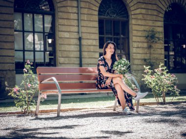 Tahta bir bankta, yaz günü elinde bir buket çiçekle dinlenen çekici bir kadının yan görünüşü.