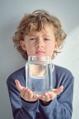 Kıvırcık sarı saçlı, kahverengi gözlü mavi süveter giyen, cam şeffaf temiz su şişesini gösteren ve beyaz arka planda kameraya bakan sevimli küçük çocuk.