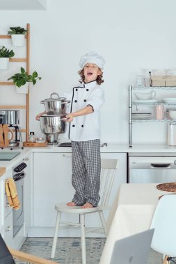 Şefin şapkası ve takım elbisesi içinde elinde tencerelerle mutfakta yemek pişirirken sandalyenin üzerinde duran sevimli, neşeli, çocuksu bir çocuk.