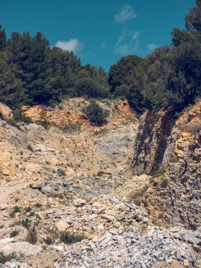 Güneşli bir günde mavi gökyüzünün altında yeşil yemyeşil bir orman bulunan kayalık yamaçta dağınık, pürüzlü taşlar.