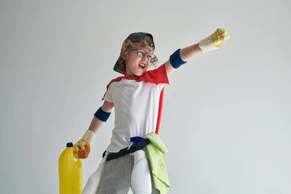 掃除用品で家庭のルーチンのための準備をしながら上昇腕で立っていると灰色の背景に対して飛んでふりを赤いケープで肯定的な男の子 — ストック写真