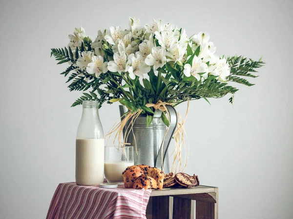 Still life of white flowers with delicate petals in metal watering can placed near glass bottle of milk and muffins in studio