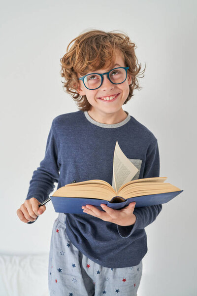 Positive boy in casual outfit and glasses holding open book and looking up while standing on white background