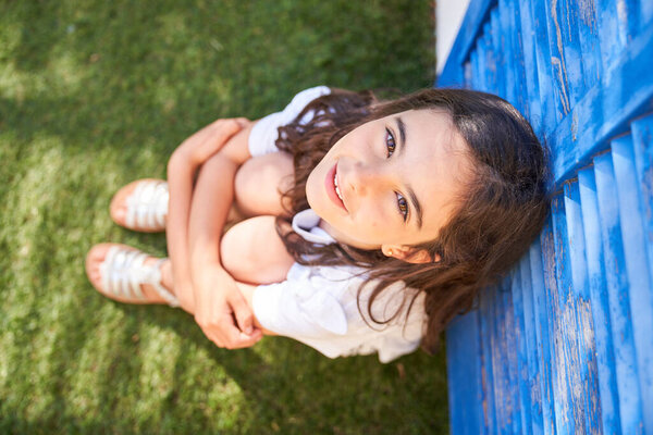 Top view of happy adorable girl looking at camera and sitting on green grass in park during summer weekend