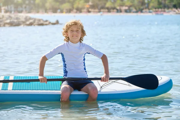 正の子供とパドル水の中に足でSupボードに座って 夏休み中に波打つ海の中で笑顔 — ストック写真