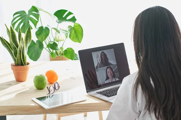 バックビューアジアの女性医師は 現代のクリニックで作業しながら テーブルでラップトップを介して患者とビデオ通話を持っている — ストック写真
