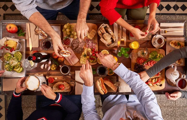 Top view group of crop faceless friends eating delicious snacks while gathering at wooden table with beverages in light kitchen