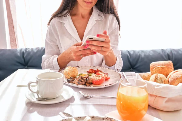 一个不愿透露姓名的女人坐在桌旁 一边吃早餐 一边吃鸡蛋 一边招手 蔬菜和橙汁 一边在智能手机上喝着新鲜咖啡 — 图库照片