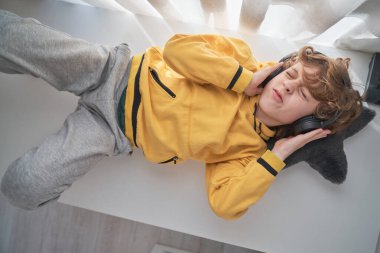 Sarı kapüşonlu ve kulaklıklı gencin evdeki masada yatıp en sevdiği müziği dinlemesi.