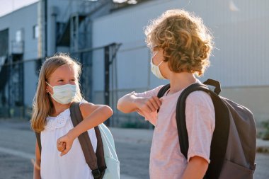 Erkek ve kız günlük giysiler ve tıbbi maskeler takmış, COVID 19 salgını sırasında şehir sokaklarında dirseklerle birbirlerini selamlıyorlar.