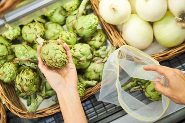 Olgun enginarı seçen ve marketten alışveriş yaparken çevre dostu kullanılabilir çanta takan yüksek açılı isimsiz bir müşteri.