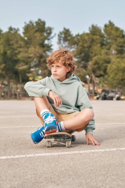 Havalı genç çocuk asfalt yolda kaykayın üzerinde oturuyor ve haftasonunun tadını çıkarırken gözlerini kaçırıyor.