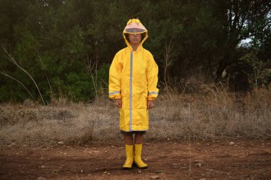 Çamurlu yolda duran ve yağmurlu sonbahar günlerinde kameraya bakan su geçirmez yağmurluk ve sarı lastik çizmeli ciddi bir çocuk.
