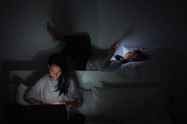 Anne yatağında oturup dizüstü bilgisayarda film izlerken çocuk evdeki karanlık odada cep telefonunu karıştırıyor.