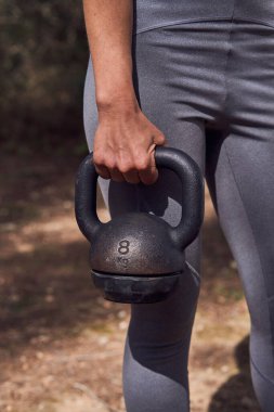 Aktif giyimli kimliği belirsiz bayan sporcu yaz parkında antrenman yaparken elinde ağır bir kettlebell ile ayakta duruyor.