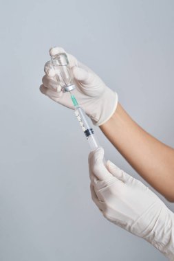 Koruyucu eldivenli tanınmayan bir doktorun elleri aşıyı şişeden şırıngaya, beyaz arka planla eşleştiriyor.