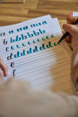 Mavi keçeli kalemle ahşap masaya yerleştirilmiş çizgili kâğıt üzerine yazı yazarken anonim sanatçının öğrendiği harflerden.