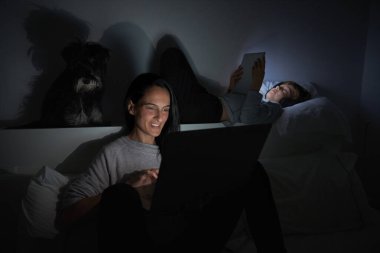 Anne gülümsüyor ve laptopta gezinirken çocuk köpek gibi yatakta uzanıyor ve evdeki karanlık odada kulaklıkla tabletten film izliyor.
