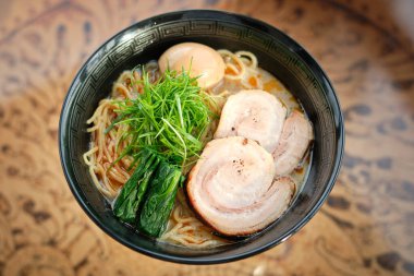 Lezzetli ramen çorbası, haşlanmış domuz yumurtası ve bulanık arka planda, masada servis edilen taze otlar.