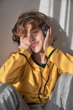 Evde kulaklıkla müzik dinlerken gülümseyen ve kameraya bakan sıradan giyinen pozitif çocuk.