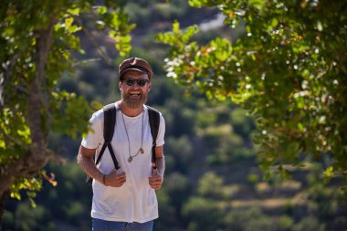 Sırt çantalı ve yuvarlak güneş gözlüklü, sakallı turist yeşil ağaçların arasında dağların bulanık arka planına karşı duruyor.