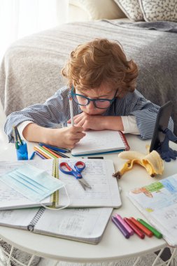 Coronavirus karantinası sırasında evde maskeyle ödev yaparken yerde oturup notlar yazan gözlüklü yakışıklı bir çocuk.