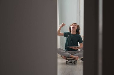 Bacak bacak üstüne atmış, kollarını kaldırmış, kaykayın üzerinde oturmuş, Tablet 'teki turnuvayı izlerken çığlık atan heyecanlı bir çocuk.