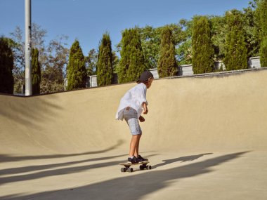 Güneşli bir günde yeşil ağaçlarla kaplı parkta asfalt yolda kaykay süren günlük kıyafetlerle ve spor ayakkabılarla kaykaycı oğlanın yan görüntüsü