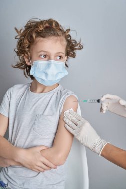Eldivenli ekin hemşiresi klinikteki beyaz duvara karşı maske takan kıvırcık saçlı çocuğa COVID 19 aşısı yapıyor.
