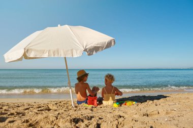 Güneşli bir yaz gününde kumlu sahilde oturup el sallayan tanınmamış küçük çocukların arka manzarası.