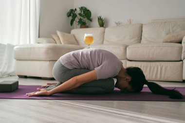 Balasana duruşunu uygulayan, yoga seansı sırasında sırtında bir bardak birayla tüm vücudu gören barışçıl kadın.