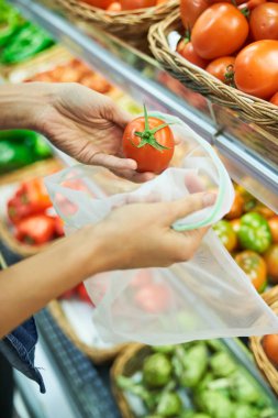 ECO dostu naylon poşete olgun kırmızı domates koyarak süpermarkette çeşitli sebzelerle tezgahın yanında duran yüzü olmayan bir alıcı.