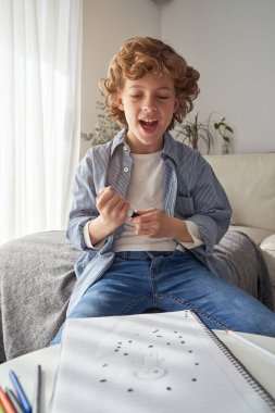 Tatmin olmuş kıvırcık saçlı, gülümseyen ve evde kanepede oturmuş sanat eserlerine bakan bir çocuk.