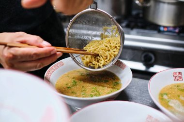Mutfaktaki masada ramen çorbası servis ederken yemek çubuklarıyla erişteyi kaseye koyan isimsiz bir aşçı.