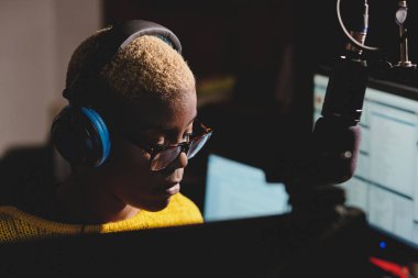 Profesyonel Afro-Amerikan radyo sunucusu kulaklıklı kadın monitörlerle masaya oturuyor ve modern yayın stüdyosunda çalışırken aşağıya bakıyor.