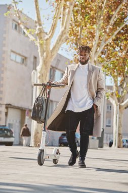 Sıcak bir ceketle gezen, güneşli şehir caddesinde mobiletiyle gezen ve kameraya bakan siyah bir erkek.