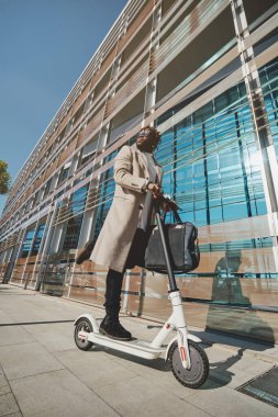 Afrika kökenli, sakallı, kıvırcık saçlı, güneş gözlüklü, bir bacağı kıvrımlı, modern binanın yakınına bakarken bir bacağı katlanmış bir erkek.