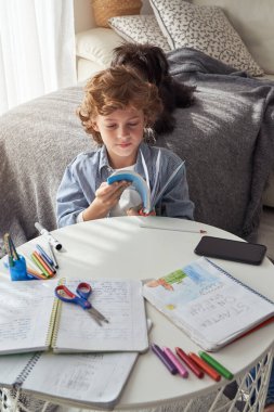 Kıvırcık elbiseli çocuk evde ödevlerini yaparken odada oturup ilginç kitaplar okuyor.