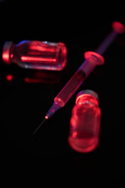 Tam tıbbi şırınga seçici odak ve kırmızı neon aydınlatmada siyah zemin üzerinde koronavirüs aşısı ile bulanık şişeler