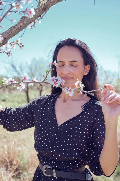 夏の田舎に立っている間 アーモンドの木の枝に咲く花に触れる目を閉じた魅力的なヒスパニックの女性 — ストック写真