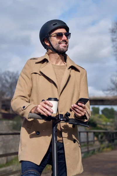 公园里 男人戴着头盔 穿着外套 手里拿着外卖咖啡 一边站在电动车上一边浏览手机 一边心满意足 — 图库照片