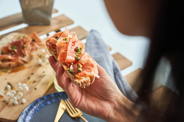 Crop Anonymous Female Eating Appetizing Bruschetta Prosciutto Capers Sitting Wooden — Stock Photo, Image